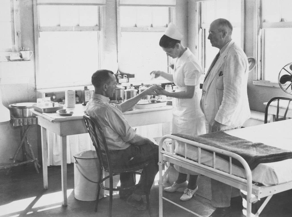 Imagen en blanco y negro de un hospital antiguo donde una enfermera y un médico atienden a un paciente en una silla